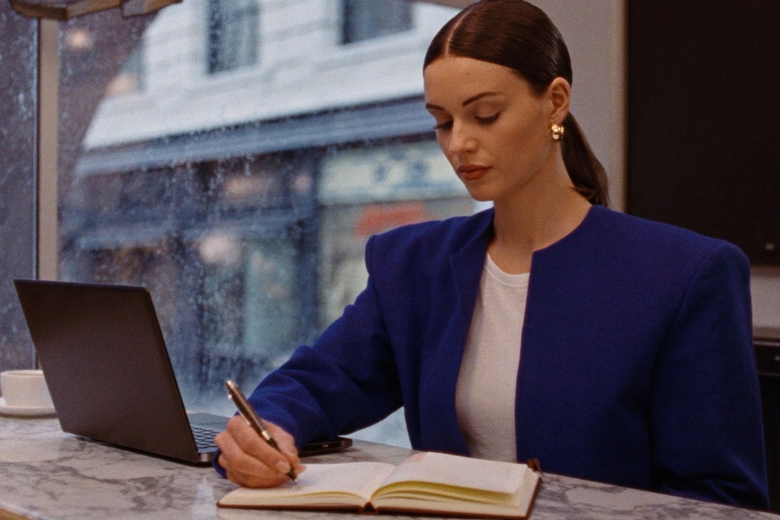 Drafting longhand with a fountain pen at a marble cafe counter, in a cobalt blazer, rain streaking the window behind