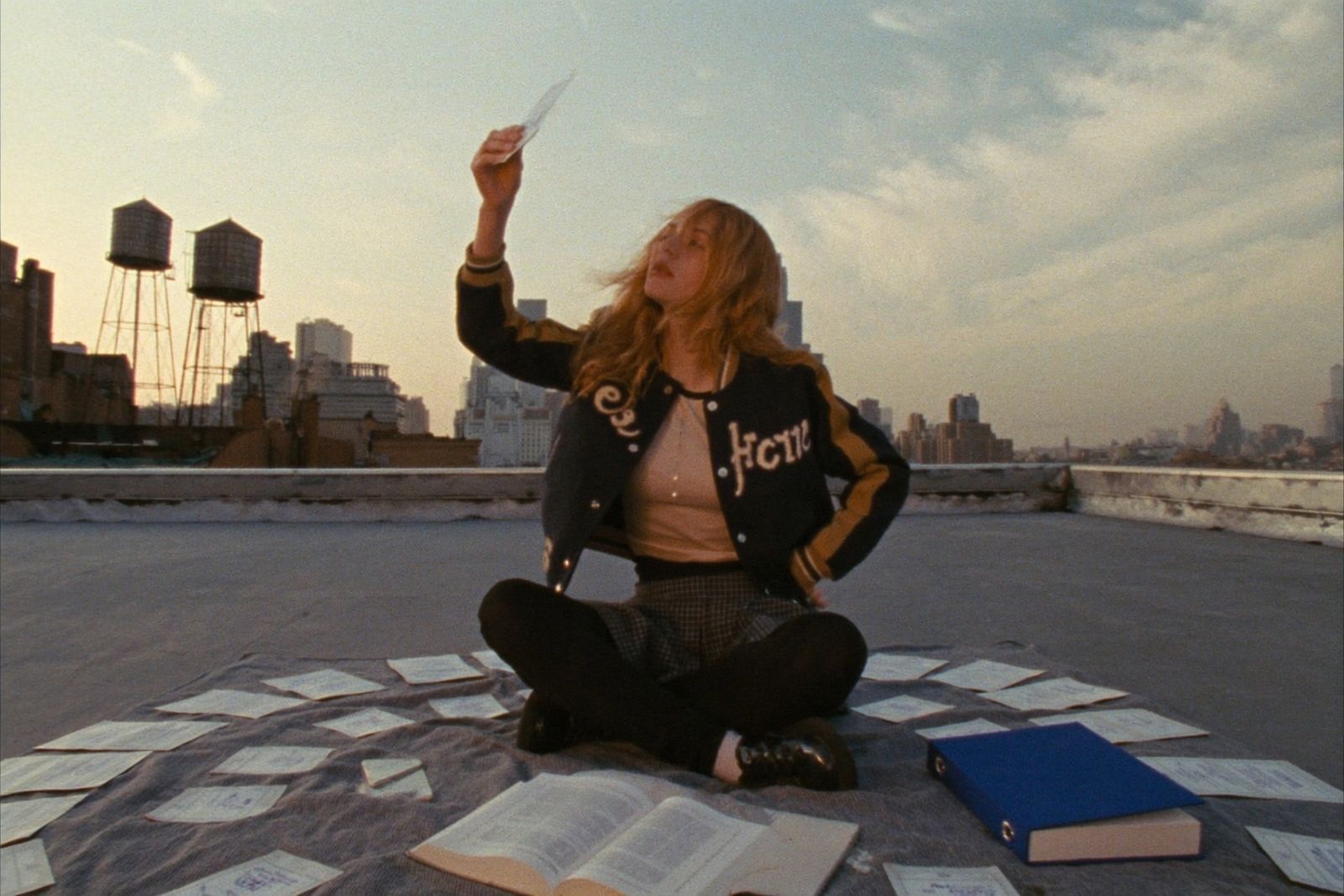 Student flipping through handwritten index cards from a cobalt binder on a rooftop, golden-hour city skyline behind