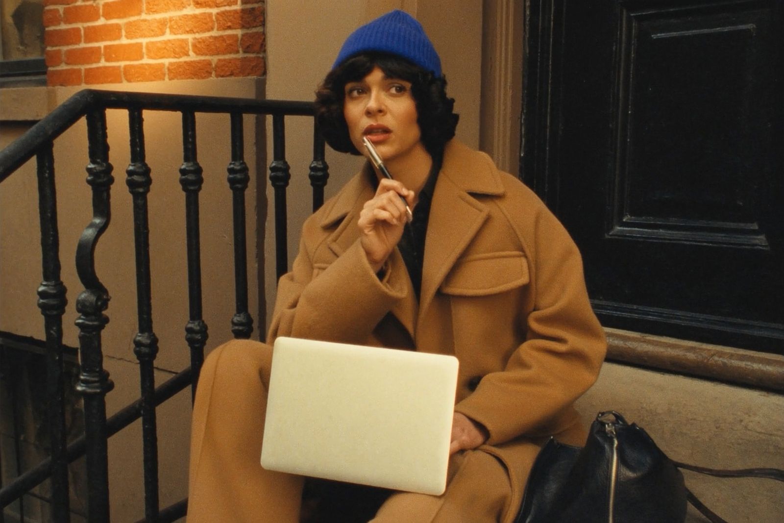 Writer sitting on a brownstone stoop in a cobalt beanie, pen to lips, laptop open