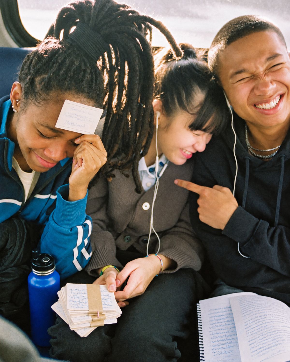 Three teens cracking up on a bus seat as one presses a flashcard to her forehead, two of them sharing wired earbuds