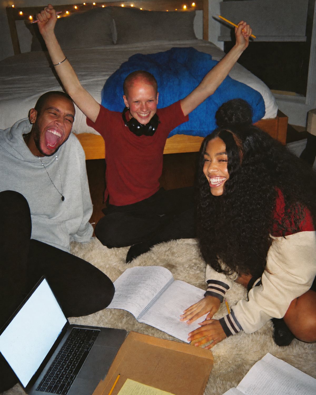 Three friends on a bedroom floor at night, one with both arms raised holding pencils, the others laughing beside a laptop and notebooks