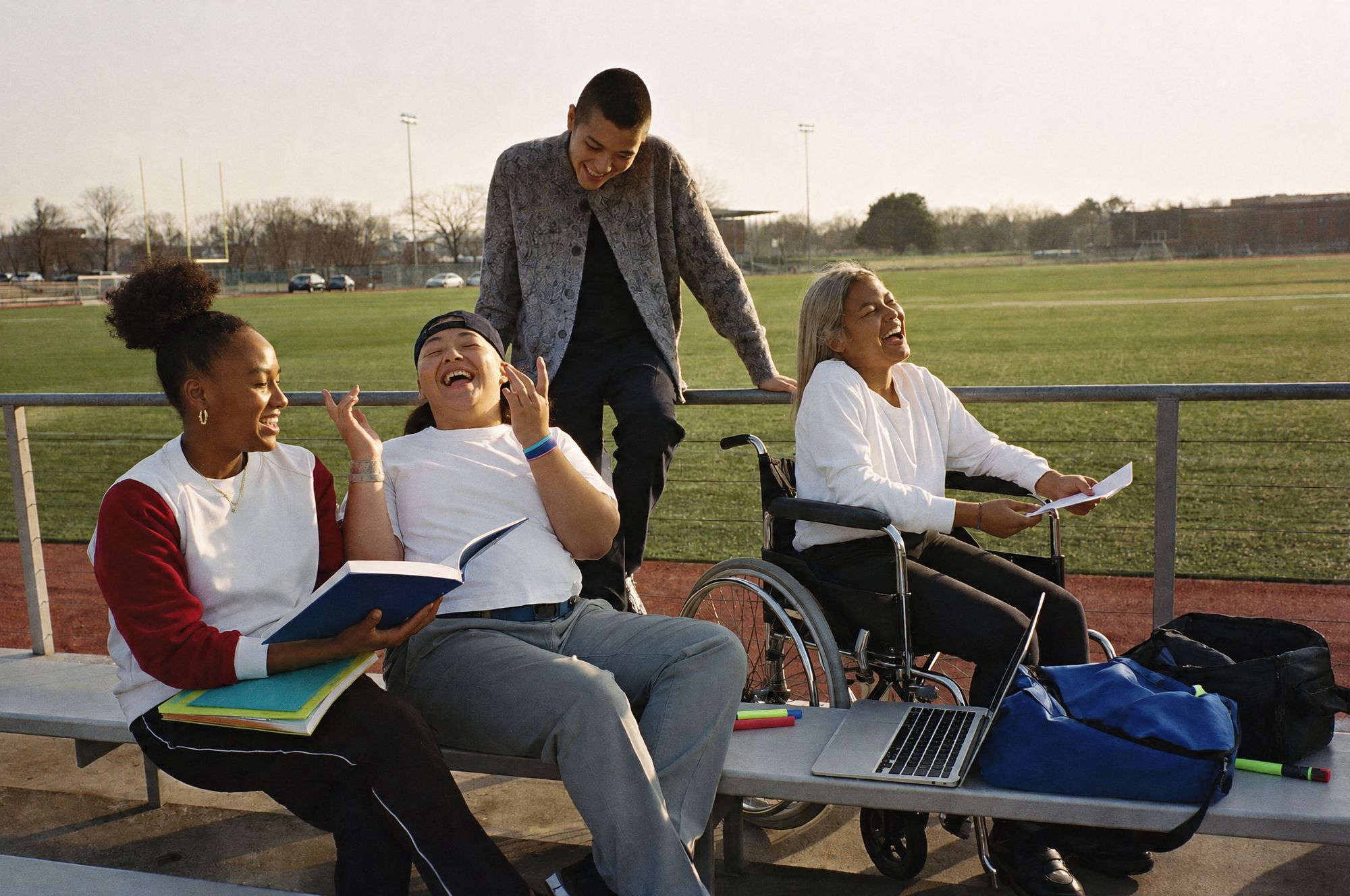 Four teens laughing on the bleachers by a sports field; one holds an open book and another, in a wheelchair, holds notes beside a laptop